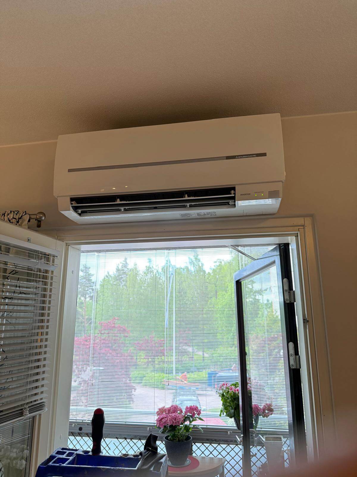 Huoneessa on ikkuna, jonka yläpuolella on valkoinen Mitsubishi Electric -ilmastointilaite. Ikkunasta näkyy puutarha, jossa on vihreitä ja punaisia puita. Ikkunalaudalla on kaksi ruukkukukkaa.