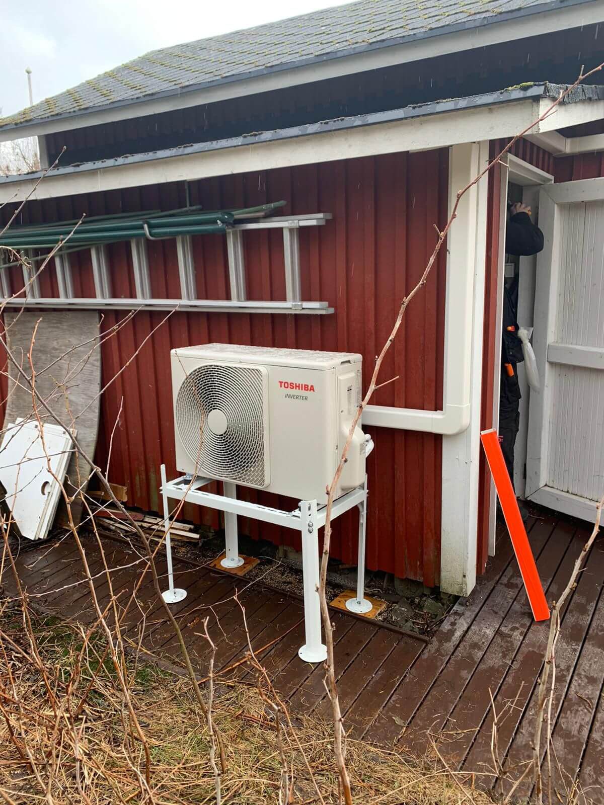 Ulkona on Toshiba-ilmastointilaite valkoisella jalustalla punaisen rakennuksen vieressä, jossa on tikkaat ja henkilö oviaukossa.