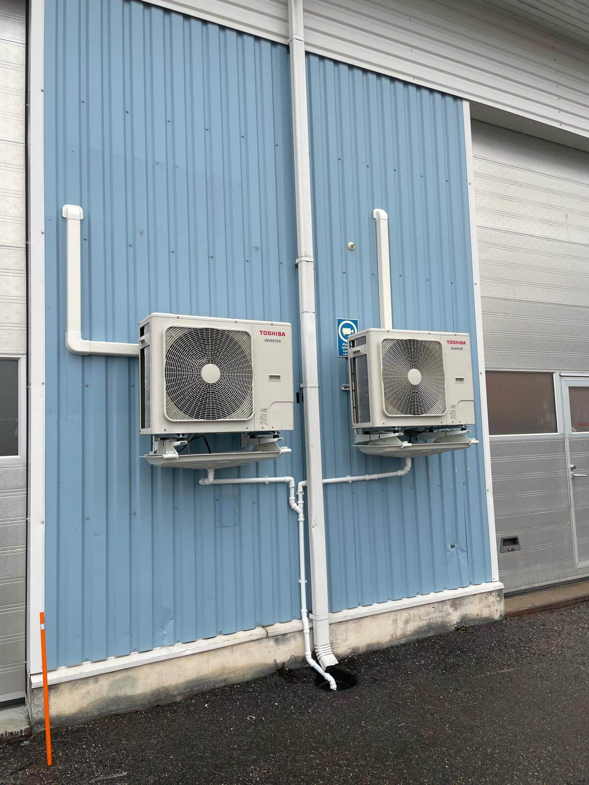 Kaksi Toshiba-ilmastointilaitetta asennettuna siniselle metalliseinälle, valkoiset putket ja sadevesikouru, ulkona päivänvalossa.