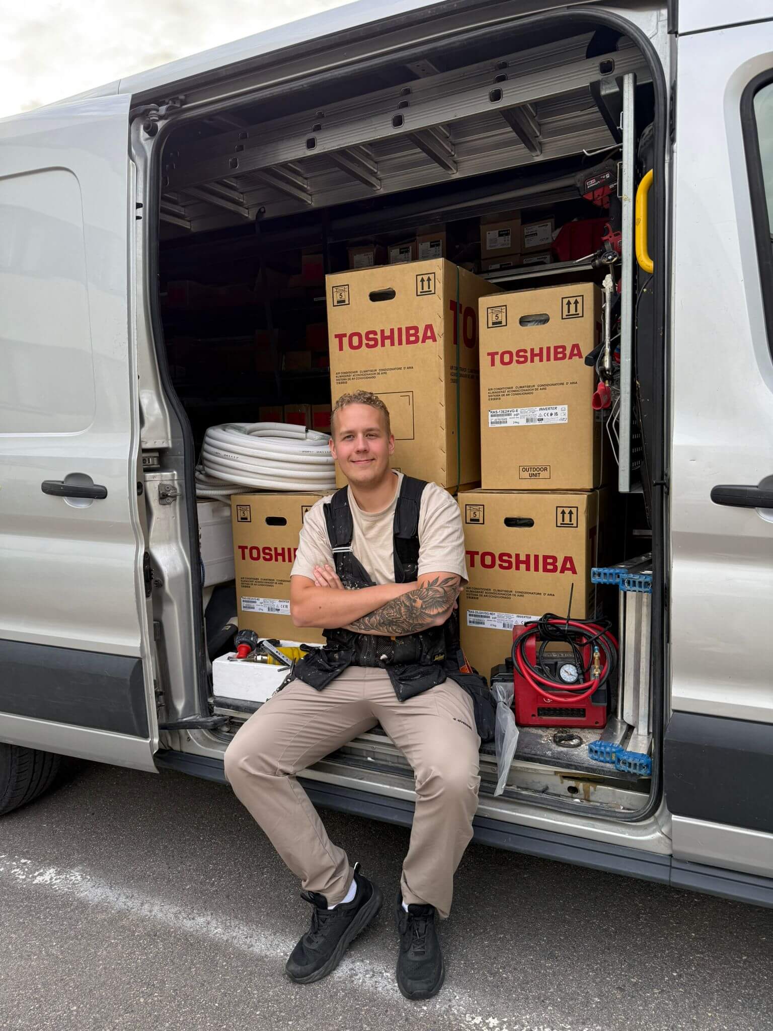 Kuva näyttää nuoren miehen istumassa pakettiauton avoimessa oviaukossa. Hänellä on yllään työhousut, t-paita ja työliivi, ja hänellä on tatuointeja käsivarsissaan. Pakettiauto on täynnä Toshiba-ilmastointilaitteiden laatikoita ja muita työkaluja. Valo on luonnollinen ja hieman pilvinen.