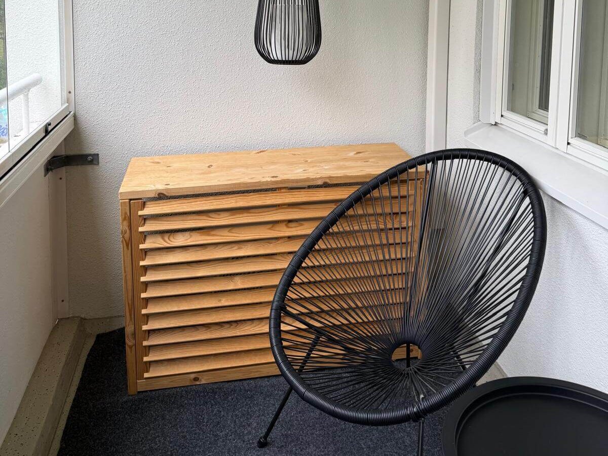 Parvekkeella on puinen säilytyslaatikko, musta roikkuva valaisin ja musta tuoli pienen pöydän vieressä.