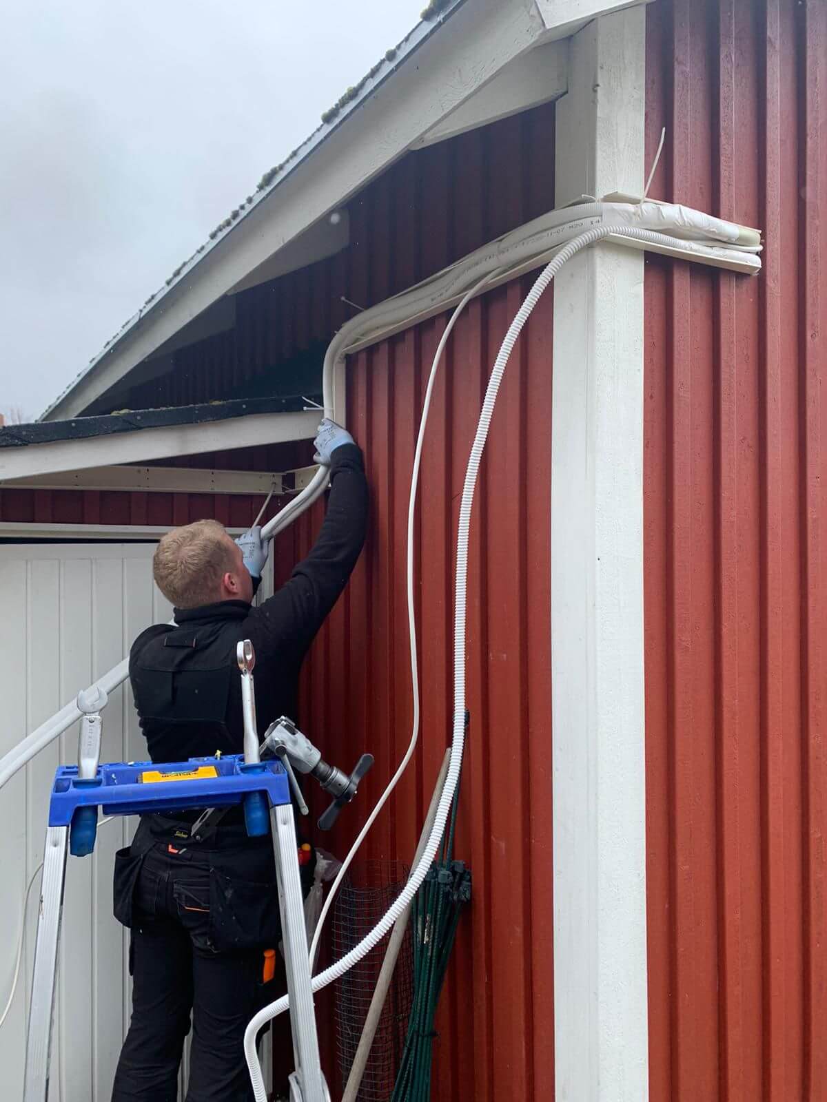 Mies asentamassa valkoista putkea punaisen rakennuksen seinään, käyttäen tikkaita ja työkaluja.