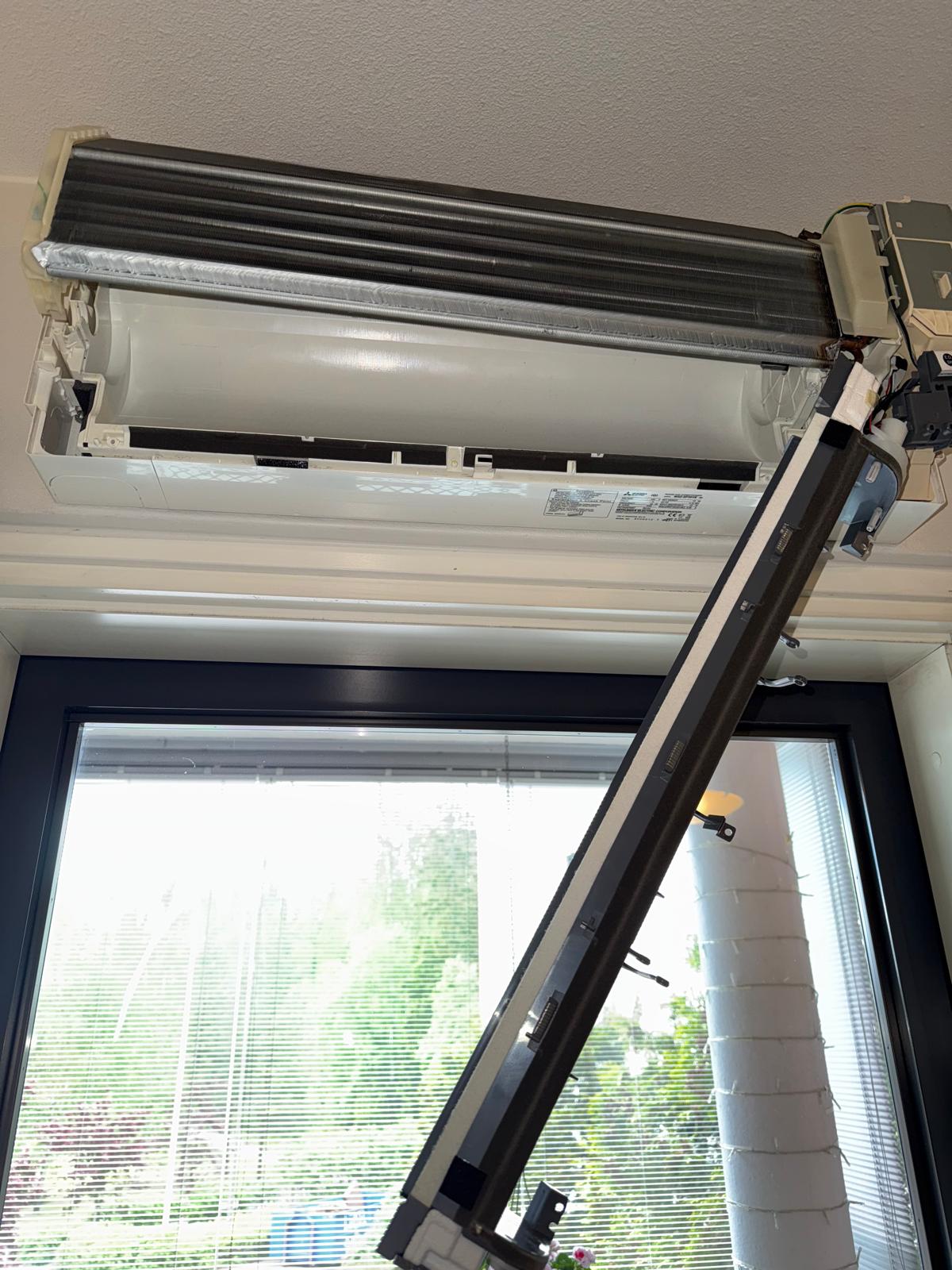 Kuva näyttää rikkoutuneen ilmastointilaitteen sisäyksikön, joka on asennettu ikkunan yläpuolelle. Laite on osittain purettu, ja sen sisäosat ovat näkyvissä. Ikkunan takana näkyy vihreää kasvillisuutta.