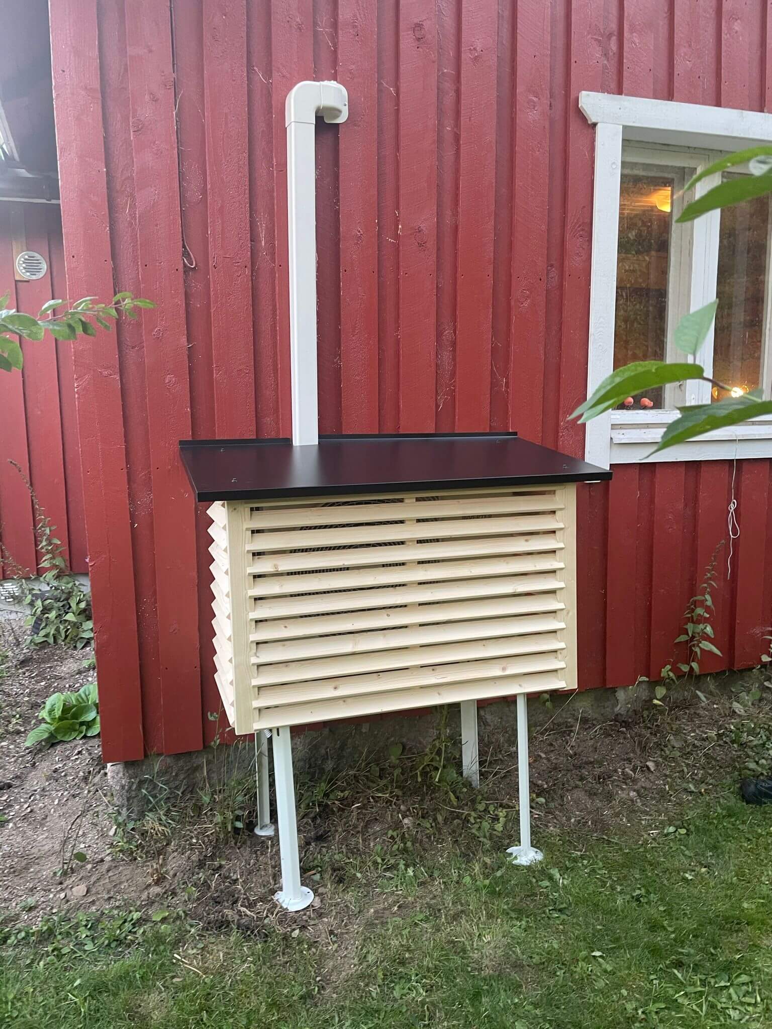 Punaisen puutalon vieressä on puinen sääsuoja, jossa on musta katto ja valkoiset jalat.