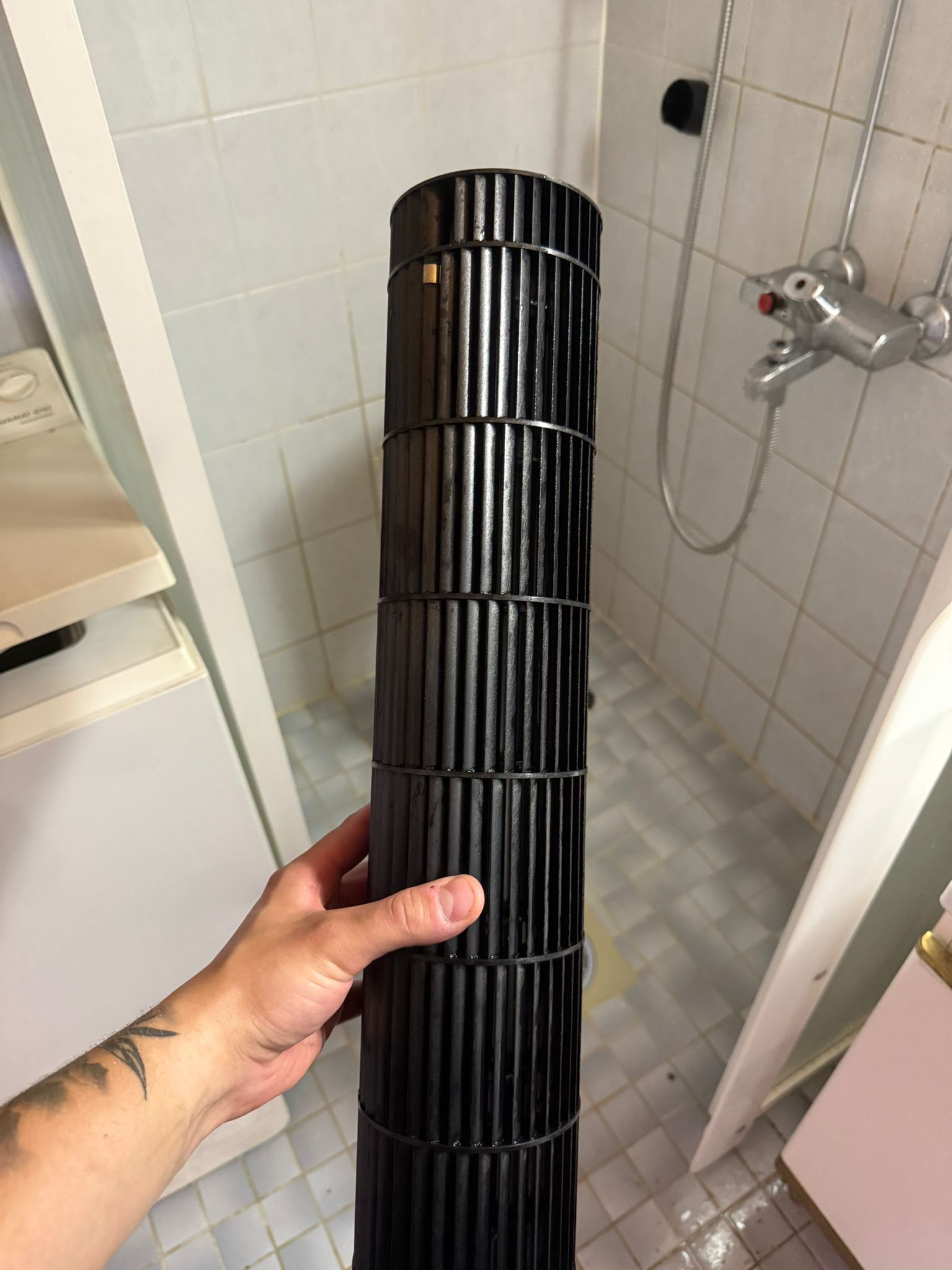 Käsivarrella pidetään mustaa, sylinterimäistä tuulettimen osaa kylpyhuoneessa, jossa on valkoiset laatat ja pesukone.